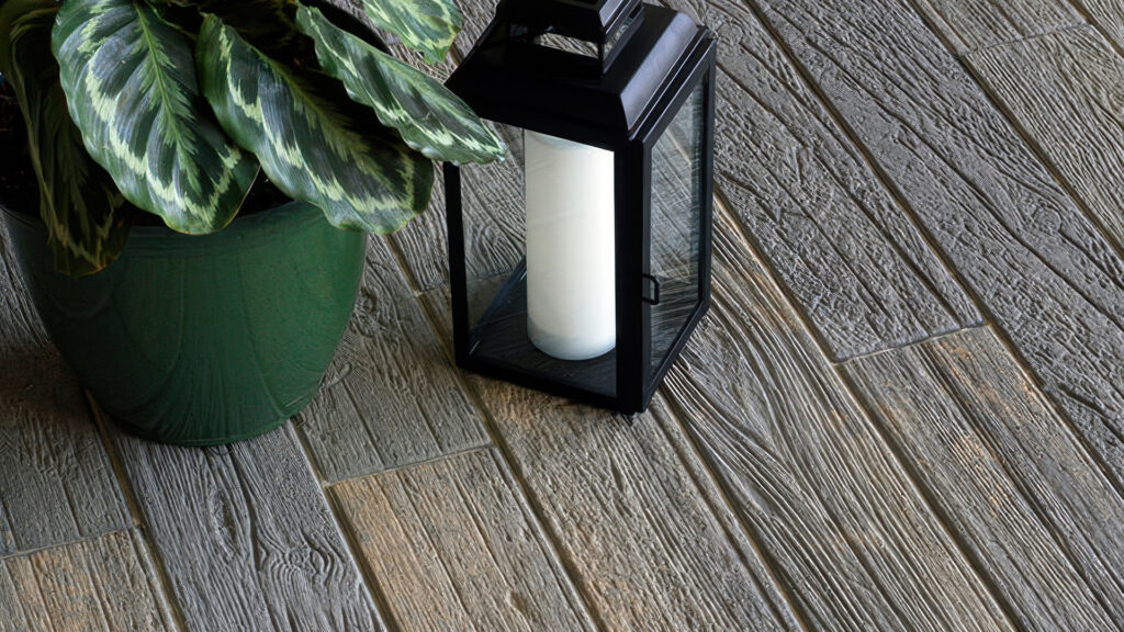 Close-up view of Timberline pavers in Dark Gray-Copper-Charcoal, showcasing a realistic wood grain texture. The pavers are arranged in a staggered pattern next to a green potted plant and a black metal lantern with a white candle inside.