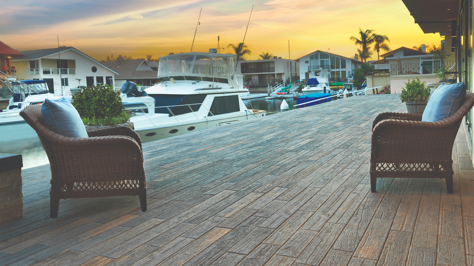 Cozy waterfront patio featuring two wicker chairs with blue cushions overlooking a calm marina lined with boats. The surface is paved with Timberline pavers in Dark Gray-Copper-Charcoal, installed in a random runner pattern that mimics natural wood grain. A golden sunset casts warm tones across the modern coastal homes and boats in the background, creating a serene, relaxing atmosphere.
