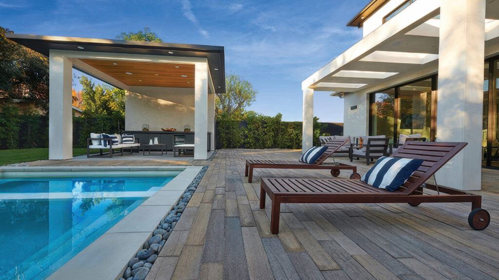 Contemporary backyard poolside patio featuring Timberline pavers in Stone-Adobe-Mocha 70mm, installed in a random running bond pattern. The space includes modern lounge chairs with striped cushions, a pebble border along the pool, and two shaded seating areas with outdoor furniture, set against a backdrop of greenery and a clear blue sky.
