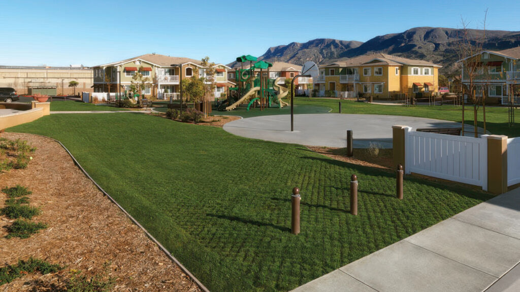 A landscaped fire lane access area featuring Gray Turfstone pavers filled with natural grass, seamlessly blending into the surrounding lawn. The area is situated within a residential complex, adjacent to a basketball half-court and children's playground, with two-story homes and mountain views in the background.