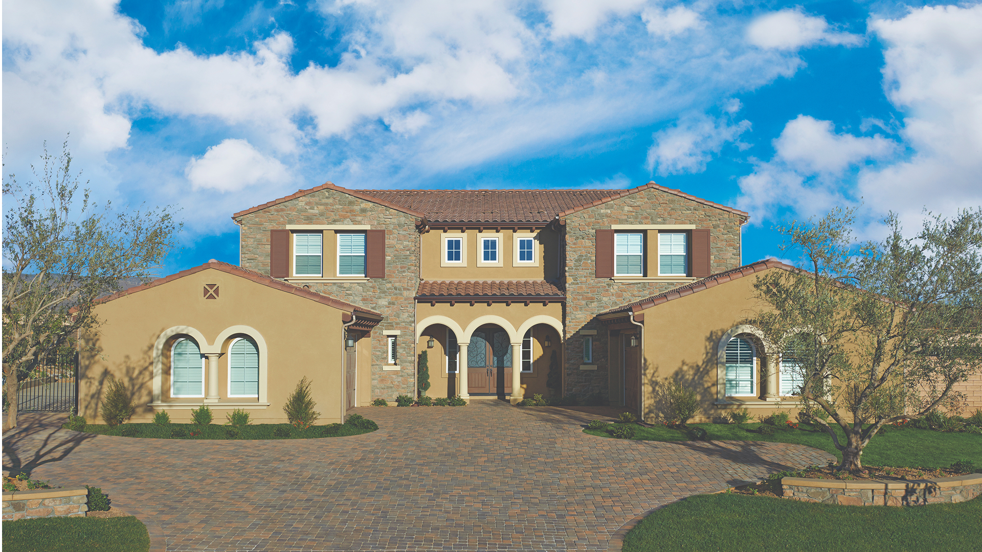 Driveway paved with Antique Cobble I and II in Tuscan blend, installed in a combination herringbone pattern. The Mediterranean-style home features arched entryways, stucco walls with stone accents, and a well-manicured front yard with olive trees and green lawn.