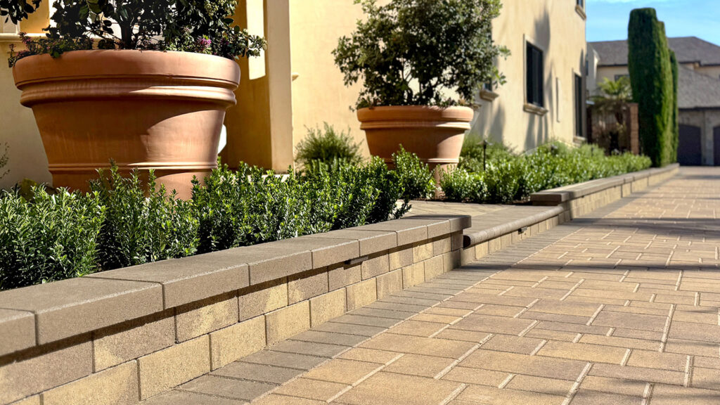 Decorative raised planter wall constructed with 12-inch Bastione Wall Stone™ in Sand-Stone-Mocha color, capped with Bastione Wall™ Cap in Mocha. The wall borders a landscaped area with neatly trimmed greenery and large terracotta planters beside a paved driveway using complementary pavers in a modern residential setting.