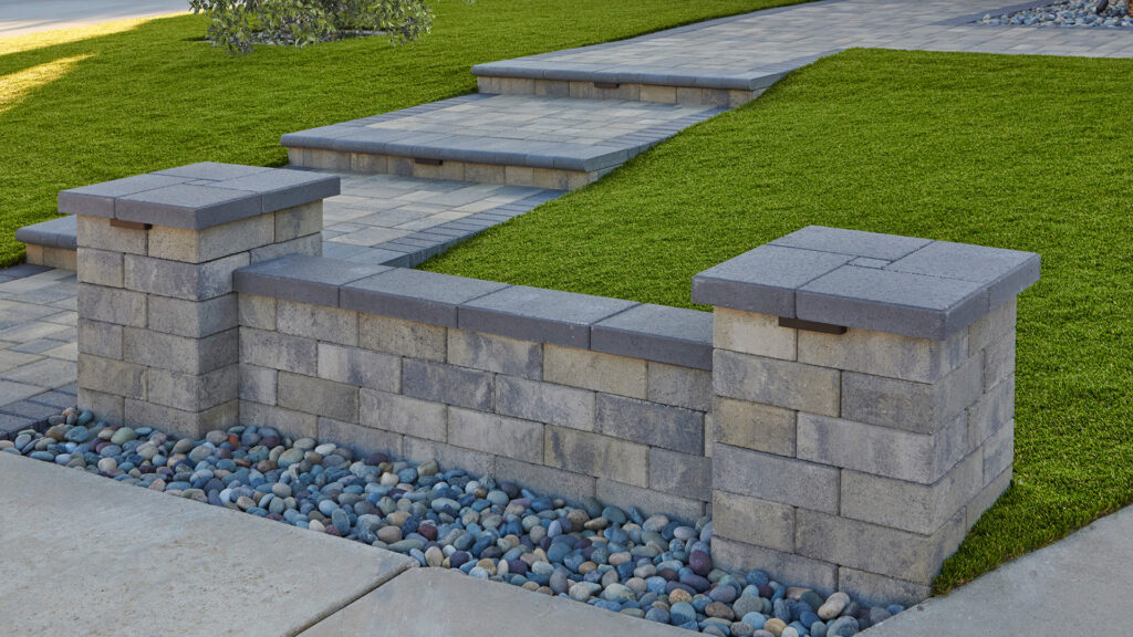 Landscape columns and retaining walls constructed with 12-inch Bastione Wall Stone™ in a Gray, Moss, and Charcoal blend, installed in a common bond pattern, frame a tiered front entry walkway. Charcoal Bastione Wall Stone™ caps top the columns and walls, while Courtyard Stone pavers in Gray, Moss, and Charcoal installed in a random ashlar pattern create the walkways and landings. Charcoal bullnose steps, integrated wall lighting, decorative river rock, and manicured lawn complete the contemporary residential landscape design.