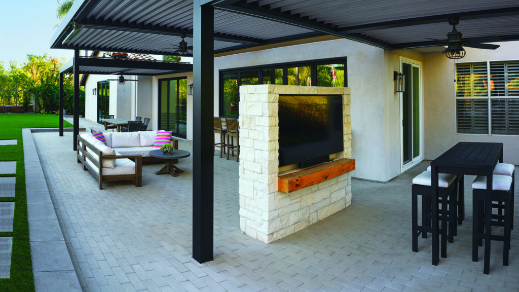 80mm 4x12 pavers in Gray, installed in a running bond pattern, create a contemporary covered patio and outdoor living area featuring lounge seating, dining spaces, a stone-clad media wall, and clean architectural lines. The paver surface complements the modern backyard design with artificial turf accents and expansive indoor-outdoor connections.