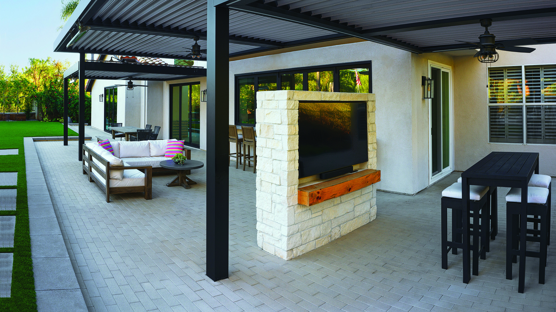 80mm 4x12 pavers in Gray, installed in a running bond pattern, create a contemporary covered patio and outdoor living area featuring lounge seating, dining spaces, a stone-clad media wall, and clean architectural lines. The paver surface complements the modern backyard design with artificial turf accents and expansive indoor-outdoor connections.