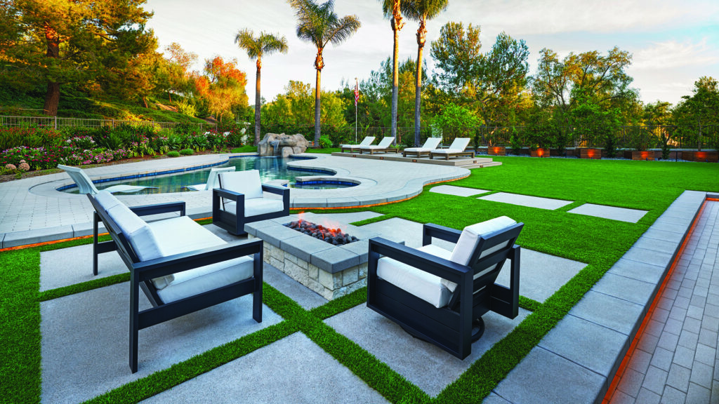 80mm 4x12 pavers in Gray, installed in a running bond pattern, form the curved pool deck and adjacent walkway in a contemporary backyard retreat. The outdoor space features a freeform swimming pool, lounge chairs, a fire pit seating area, artificial turf accents, and lush landscaping with mature trees and colorful garden beds.