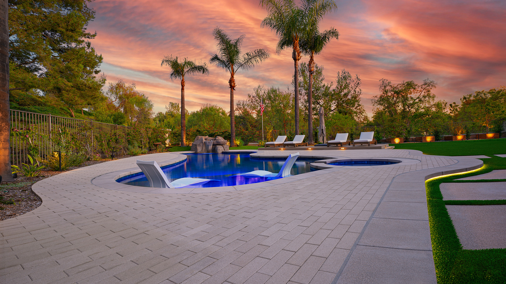 80mm 4x12 pavers in Gray, installed in a running bond pattern, create a sweeping pool deck around a freeform swimming pool and spa. The contemporary outdoor space features in-water lounge chairs, artificial turf accents, palm trees, landscape lighting, and resort-style seating areas beneath a colorful sunset sky.