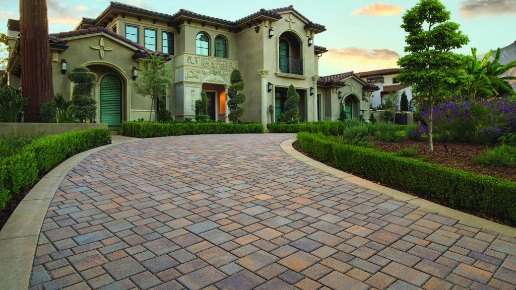 Permeable Courtyard pavers in a custom Tuscan color blend, installed in a random ashlar pattern, create a sweeping circular driveway leading to an upscale Mediterranean-style home, surrounded by manicured hedges, sculpted topiary, ornamental trees, and lush landscape plantings at sunset.