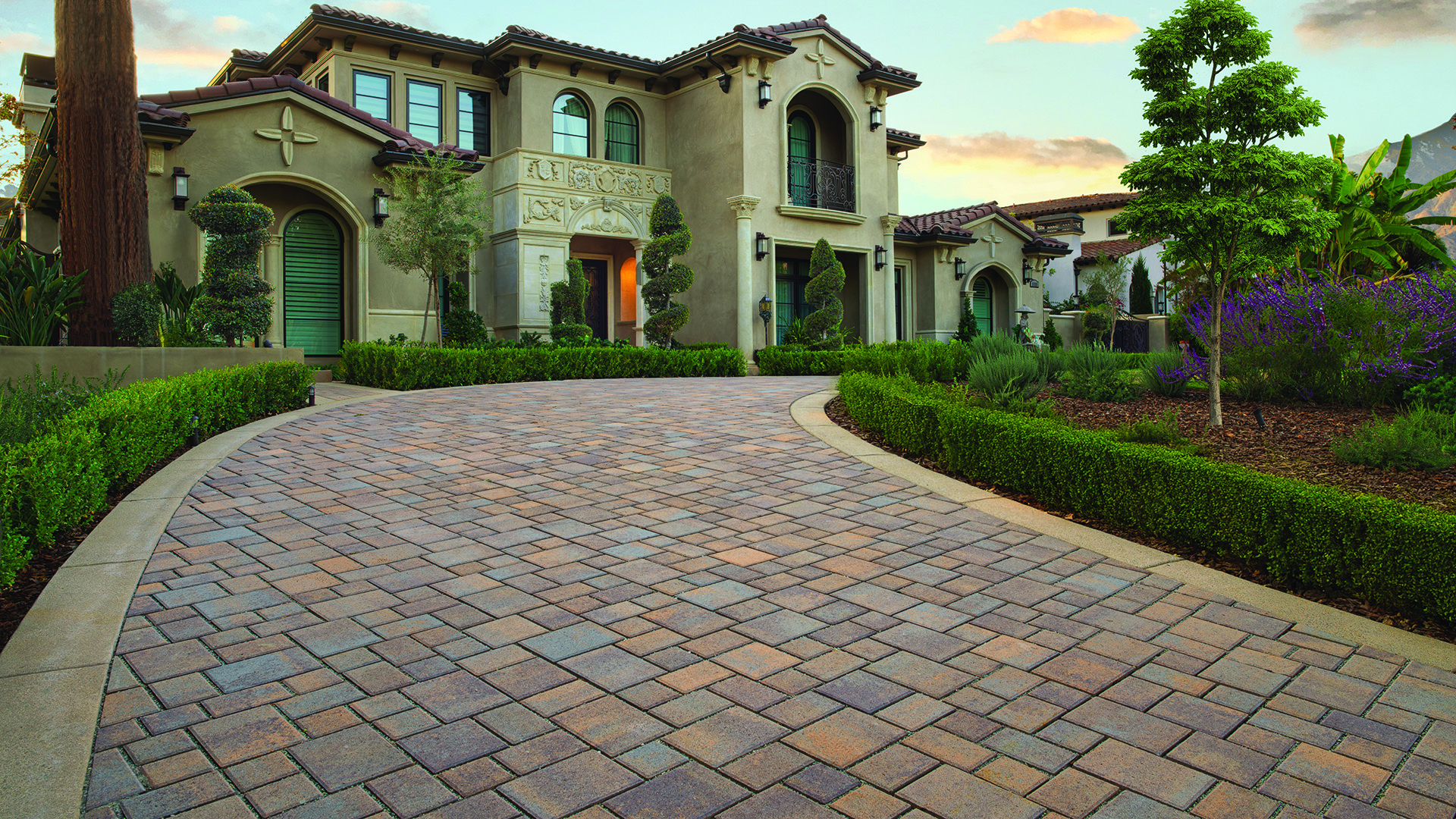 Permeable Courtyard pavers in a custom Tuscan color blend, installed in a random ashlar pattern, create a sweeping circular driveway leading to an upscale Mediterranean-style home, surrounded by manicured hedges, sculpted topiary, ornamental trees, and lush landscape plantings at sunset.