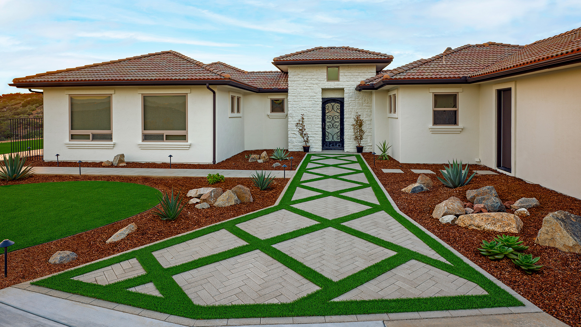 Front entry walkway featuring 80mm 4x12 pavers in Gray installed in a herringbone pattern, arranged within geometric turf inlays leading to a modern home entrance. The light gray pavers create a striking diamond design bordered by green grass, with drought-tolerant landscaping, decorative boulders, and clean architectural lines framing the entry.
