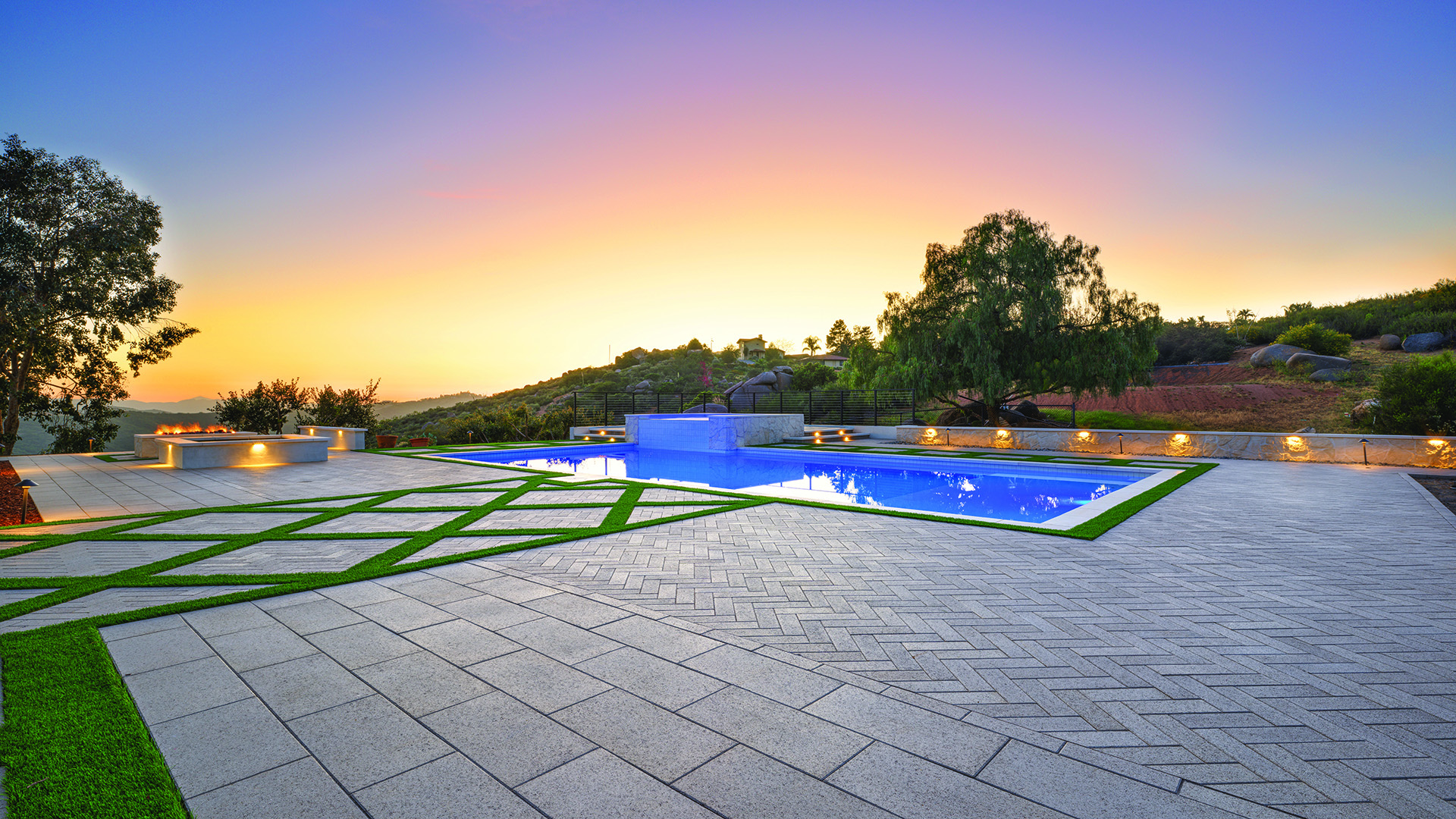 80mm 4x12 pavers in Gray, installed in a herringbone pattern, create an expansive pool deck surrounding a modern rectangular swimming pool. In the foreground, 60mm 12x24 Gray pavers installed in a running bond pattern form a geometric patio with artificial turf accents, while landscape lighting, a fire feature, and rolling hillside views enhance the contemporary outdoor setting at sunset.
