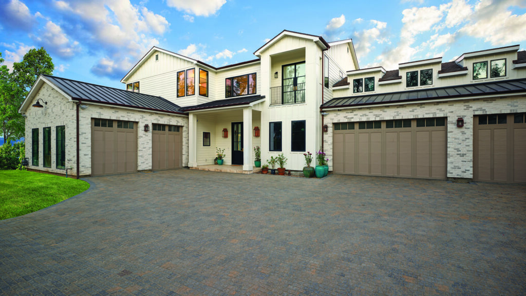 70mm Torino pavers in a dark gray, copper, and charcoal color blend, installed in a random running bond pattern, create a spacious driveway and entry court in front of a modern farmhouse-style home with white brick, dark metal roofing, and three-car garage doors beneath a dramatic evening sky.