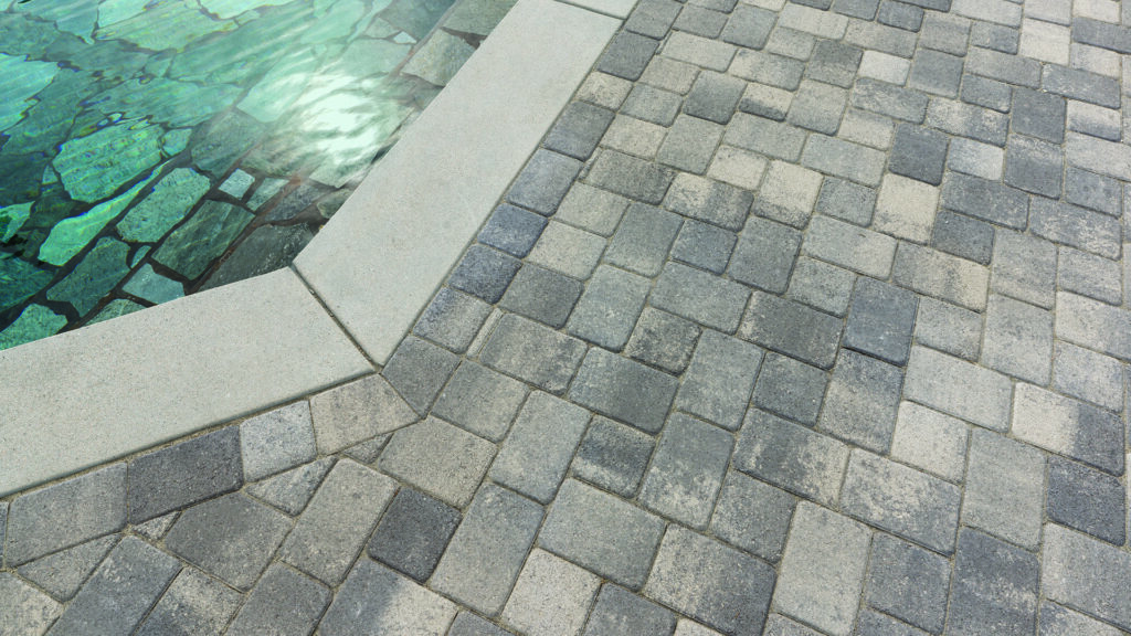 Antique Cobble I & II pavers in a gray and charcoal color blend, installed in an “I” pattern, create a textured pool deck surface adjacent to light-colored concrete coping. A border of Antique Cobble I & II in a random runner pattern frames the installation, highlighting the contrasting paver layout and color variation beside the pool.