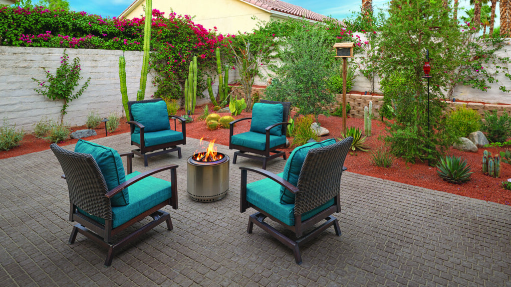 70mm Torino pavers in Mocha, installed in a random running bond pattern, create a warm and inviting patio seating area with four cushioned lounge chairs arranged around a fire pit. The outdoor space is surrounded by a desert-inspired landscape featuring cacti, succulents, flowering bougainvillea, and decorative gravel.