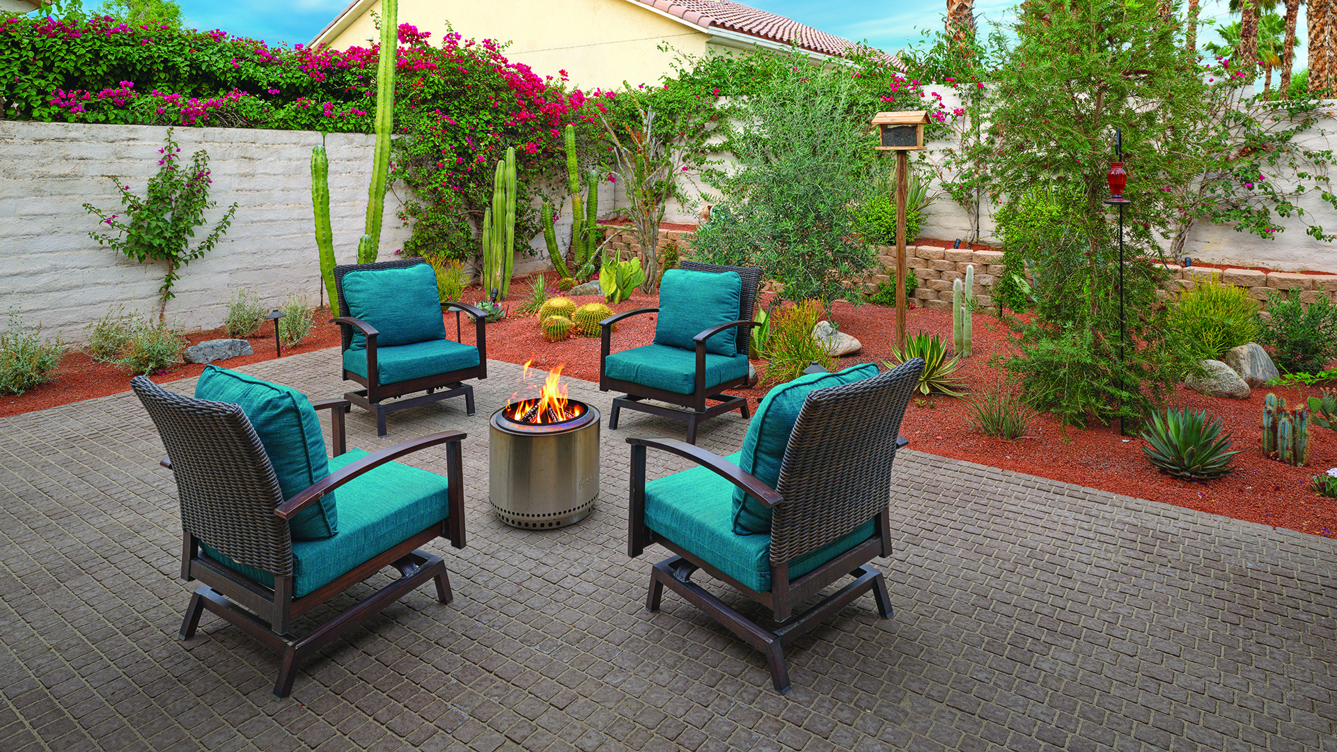 70mm Torino pavers in Mocha, installed in a random running bond pattern, create a warm and inviting patio seating area with four cushioned lounge chairs arranged around a fire pit. The outdoor space is surrounded by a desert-inspired landscape featuring cacti, succulents, flowering bougainvillea, and decorative gravel.