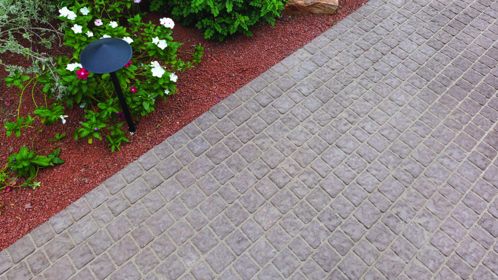 Close-up view of a garden pathway paved with 70mm Torino pavers in Mocha, installed in a random running bond pattern. The walkway is bordered by red decorative gravel, flowering plants, and landscape lighting, highlighting the textured surface and warm, natural color tones of the pavers.
