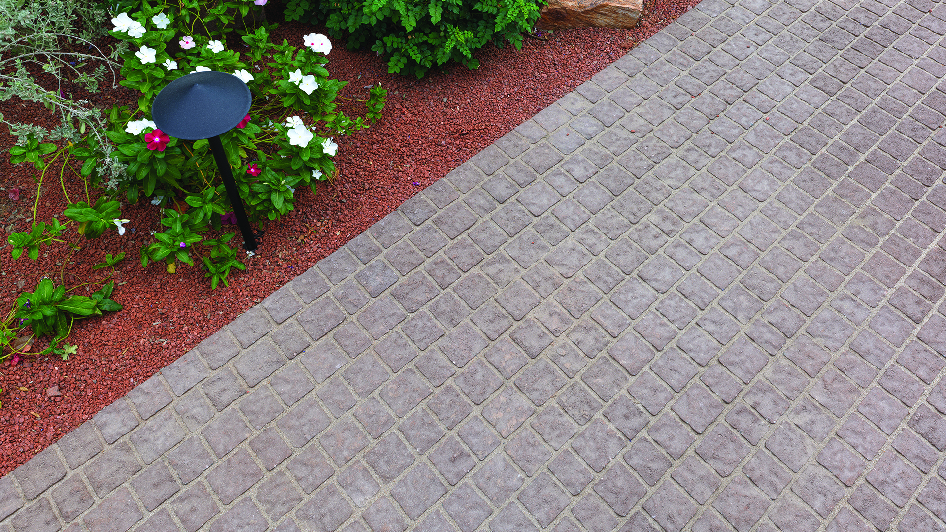 Close-up view of a garden pathway paved with 70mm Torino pavers in Mocha, installed in a random running bond pattern. The walkway is bordered by red decorative gravel, flowering plants, and landscape lighting, highlighting the textured surface and warm, natural color tones of the pavers.