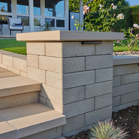 A landscape staircase and retaining wall constructed with gray Bastione Wall Stone™. The column is built with 12-inch Bastione Wall Stone™ in Gray, while the adjoining retaining wall uses 24-inch Bastione Wall Stone™ in Gray installed in a running bond pattern. Smooth concrete stair treads, integrated wall lighting, and surrounding garden plantings complete the contemporary outdoor design.