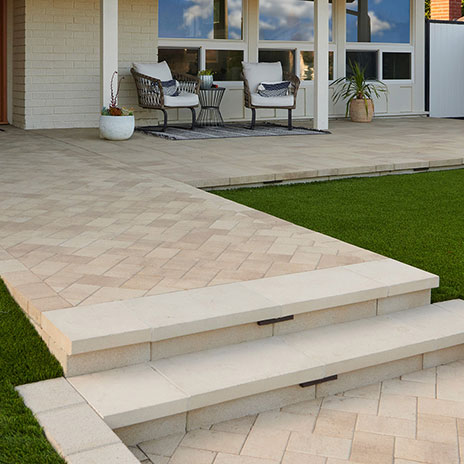 A residential front entry features Porcellana 6-inch pavers in a Light Sand, Gray, and Stone color blend, installed in a herringbone pattern along the main walkway. Adjacent patio areas use Porcellana 18-inch pavers in the same color blend, installed in a random ashlar pattern. A low retaining wall constructed with 12-inch Bastione Wall Stone™ in Gray borders the entry, while broad steps, manicured lawn, and contemporary outdoor seating create a clean and inviting approach to the home.