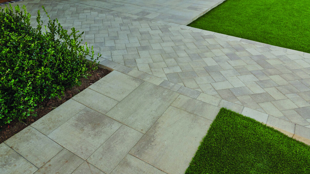 Landscape walkway showcasing Porcellana 6-inch pavers in Light Sand-Gray-Stone installed in a herringbone pattern, bordered by Porcellana 18-inch pavers in Light Sand-Gray-Stone installed in a random ashlar pattern. The light-toned pavers create a clean transition between lawn and planting beds, highlighting the contrasting paving patterns and contemporary outdoor design.