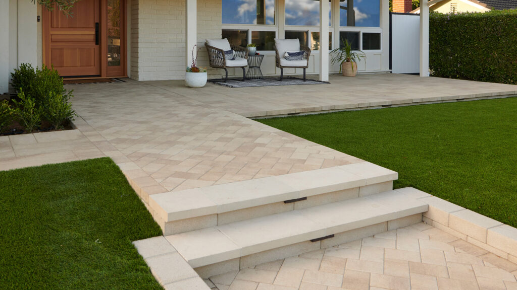 A residential front entry features Porcellana 6-inch pavers in a Light Sand, Gray, and Stone color blend, installed in a herringbone pattern along the main walkway. Adjacent patio areas use Porcellana 18-inch pavers in the same color blend, installed in a random ashlar pattern. A low retaining wall constructed with 12-inch Bastione Wall Stone™ in Gray borders the entry, while broad steps, manicured lawn, and contemporary outdoor seating create a clean and inviting approach to the home.
