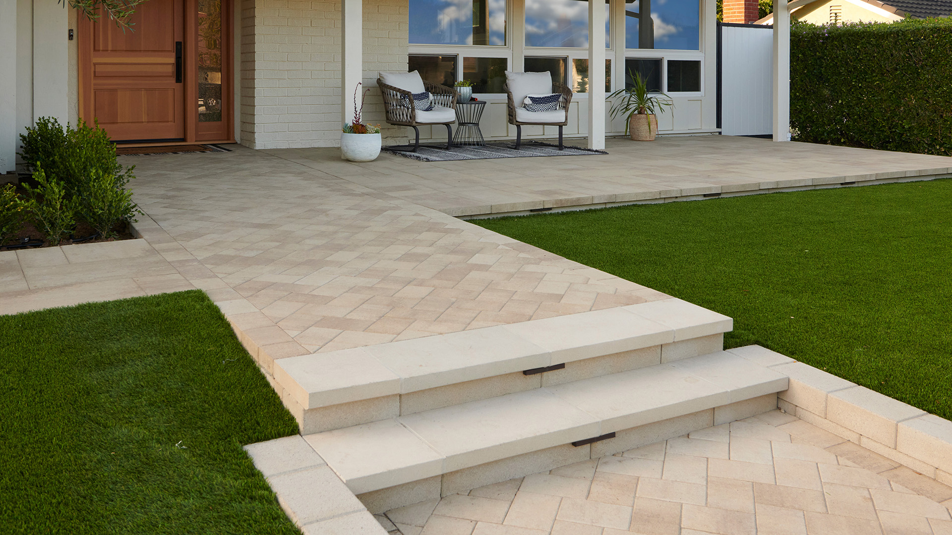 A residential front entry features Porcellana 6-inch pavers in a Light Sand, Gray, and Stone color blend, installed in a herringbone pattern along the main walkway. Adjacent patio areas use Porcellana 18-inch pavers in the same color blend, installed in a random ashlar pattern. A low retaining wall constructed with 12-inch Bastione Wall Stone™ in Gray borders the entry, while broad steps, manicured lawn, and contemporary outdoor seating create a clean and inviting approach to the home.