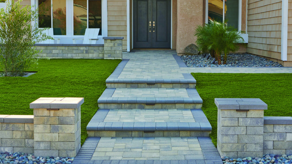 Landscape columns and retaining walls constructed with 12-inch Bastione Wall Stone™ in a Gray, Moss, and Charcoal blend, installed in a common bond pattern, frame a tiered front entry walkway leading to a residential home. Charcoal Bastione Wall Stone™ caps top the columns and walls, while Courtyard Stone pavers in Gray, Moss, and Charcoal installed in a random ashlar pattern create the walkways and landings. Charcoal bullnose steps and a Courtyard 6x9 Charcoal border provide contrast and definition, complemented by manicured lawns and decorative river rock landscaping.