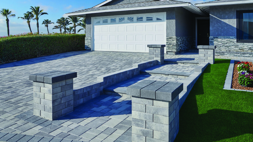 Landscape columns and retaining walls constructed with 12-inch Bastione Wall Stone™ in a Gray-Charcoal blend, installed in a common bond pattern, frame a raised entry walkway and stairway leading to a contemporary home. Charcoal Bastione Wall Stone™ caps top the columns, while Courtyard Stone pavers in Gray-Charcoal installed in a random ashlar pattern create the driveway and walkways. Charcoal bullnose steps provide a clean transition between elevations, complemented by manicured lawn and coastal landscaping.