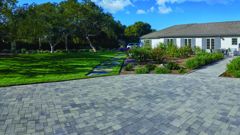 Gray and charcoal distressed Castle Cobble I & II concrete pavers installed in a random ashlar pattern create a large patio and walkway beside a white home, bordered by manicured lawn, colorful rose gardens, and mature shade trees under a bright blue sky.