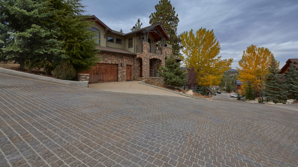 70mm Torino pavers in a custom Mocha and Charcoal color blend, installed in a random running bond pattern, create a textured residential street surface in a mountain community. The pavers complement a rustic stone-and-wood home surrounded by evergreen trees and colorful autumn foliage under an overcast sky.