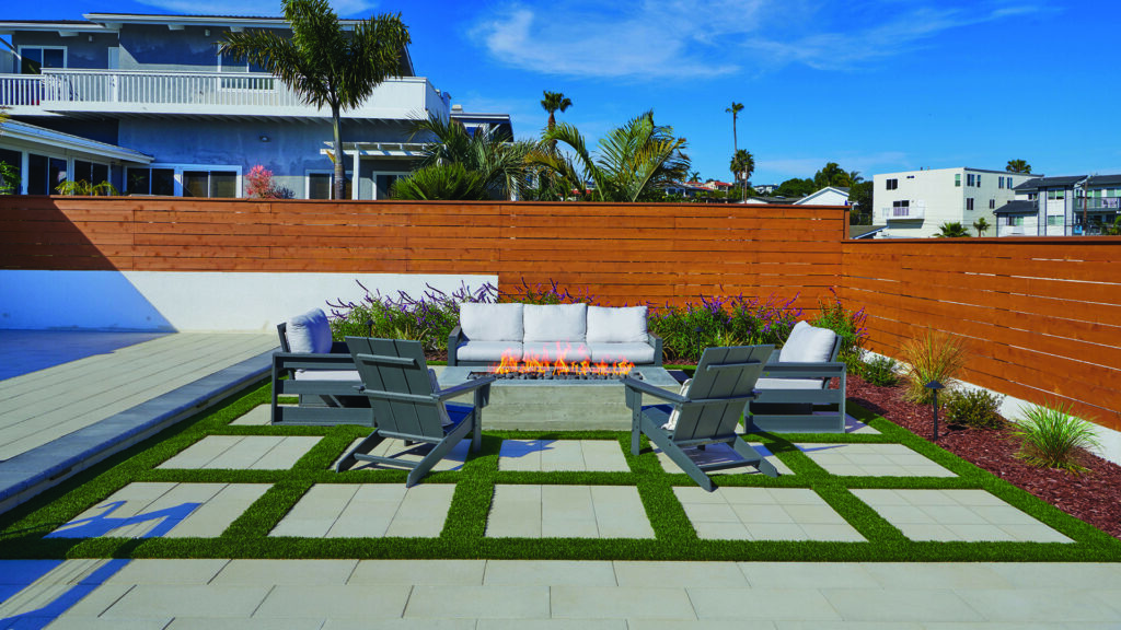 60mm 12x24 pavers in Solid Gray, installed in a running bond pattern, create a modern outdoor living space with geometric paving, artificial turf joints, and a central fire feature surrounded by lounge seating. The contemporary backyard is enclosed by a wood privacy fence and accented with palms, ornamental grasses, and colorful landscape plantings.