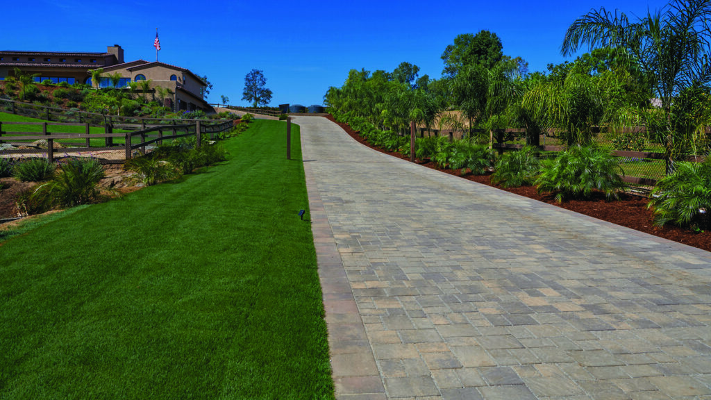Sand, stone, and mocha Permeable Slate pavers installed in a random ashlar pattern create a long residential driveway leading to a hillside estate, bordered by manicured lawns, palm trees, landscaped planting beds, and ranch-style fencing under a clear blue sky.