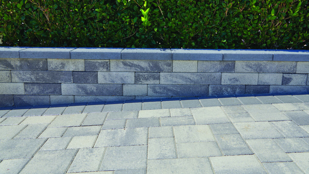 A retaining wall constructed with 3 Piece Bastione Wall Stone™ in a Gray-Charcoal blend, installed in a random runner pattern, borders a paved outdoor living area. The wall is topped with Charcoal Bastione Wall Stone™ caps, while Courtyard Stone pavers in Gray-Charcoal installed in a random ashlar pattern create the patio surface. A Courtyard 6x9 Charcoal border provides definition between the wall and paving, with a manicured hedge backdrop completing the landscape design.