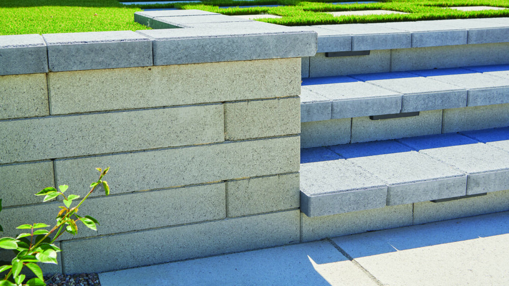 A retaining wall and tiered landscape steps constructed with 24-inch Bastione Wall Stone™ in Gray, installed in a running bond pattern. Charcoal Bastione Wall Stone™ caps finish the wall and step treads, while integrated lighting beneath the caps highlights the clean lines and contemporary design of the elevated outdoor living space.
