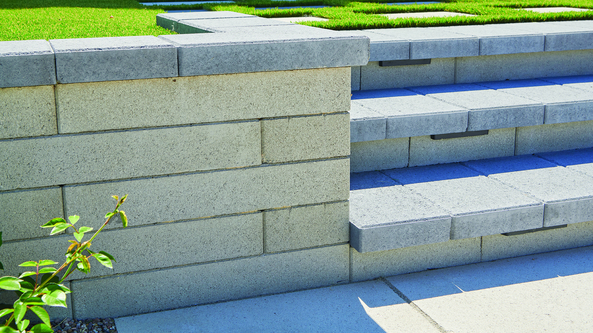A retaining wall and tiered landscape steps constructed with 24-inch Bastione Wall Stone™ in Gray, installed in a running bond pattern. Charcoal Bastione Wall Stone™ caps finish the wall and step treads, while integrated lighting beneath the caps highlights the clean lines and contemporary design of the elevated outdoor living space.