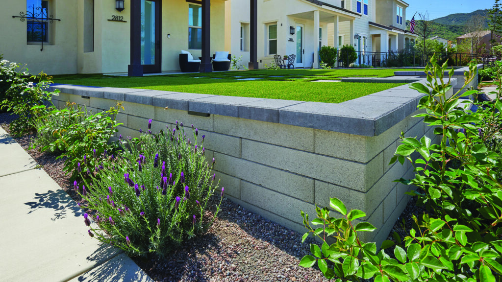 A retaining wall constructed with 24-inch Bastione Wall Stone™ in Gray, installed in a one-third running bond pattern, defines an elevated front lawn and landscape area. Charcoal Bastione Wall Stone™ caps finish the wall with clean, contemporary lines, while integrated lighting beneath the caps highlights the structure. Flowering plants, shrubs, and a modern residential home provide an attractive backdrop to the landscape design.