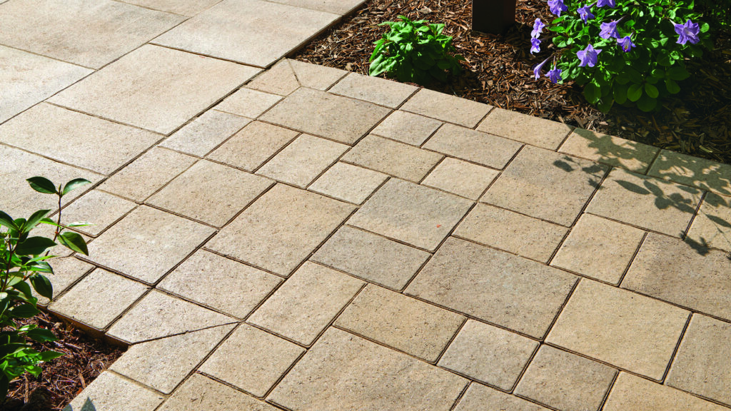 Close-up of a paved walkway featuring Porcellana 9-inch pavers in Light Sand-Gray-Stone installed in a random ashlar pattern in the foreground. Larger Porcellana 18-inch pavers in matching Light Sand-Gray-Stone are installed in a random ashlar pattern in the background, creating a coordinated transition between paving areas bordered by landscaped planting beds.