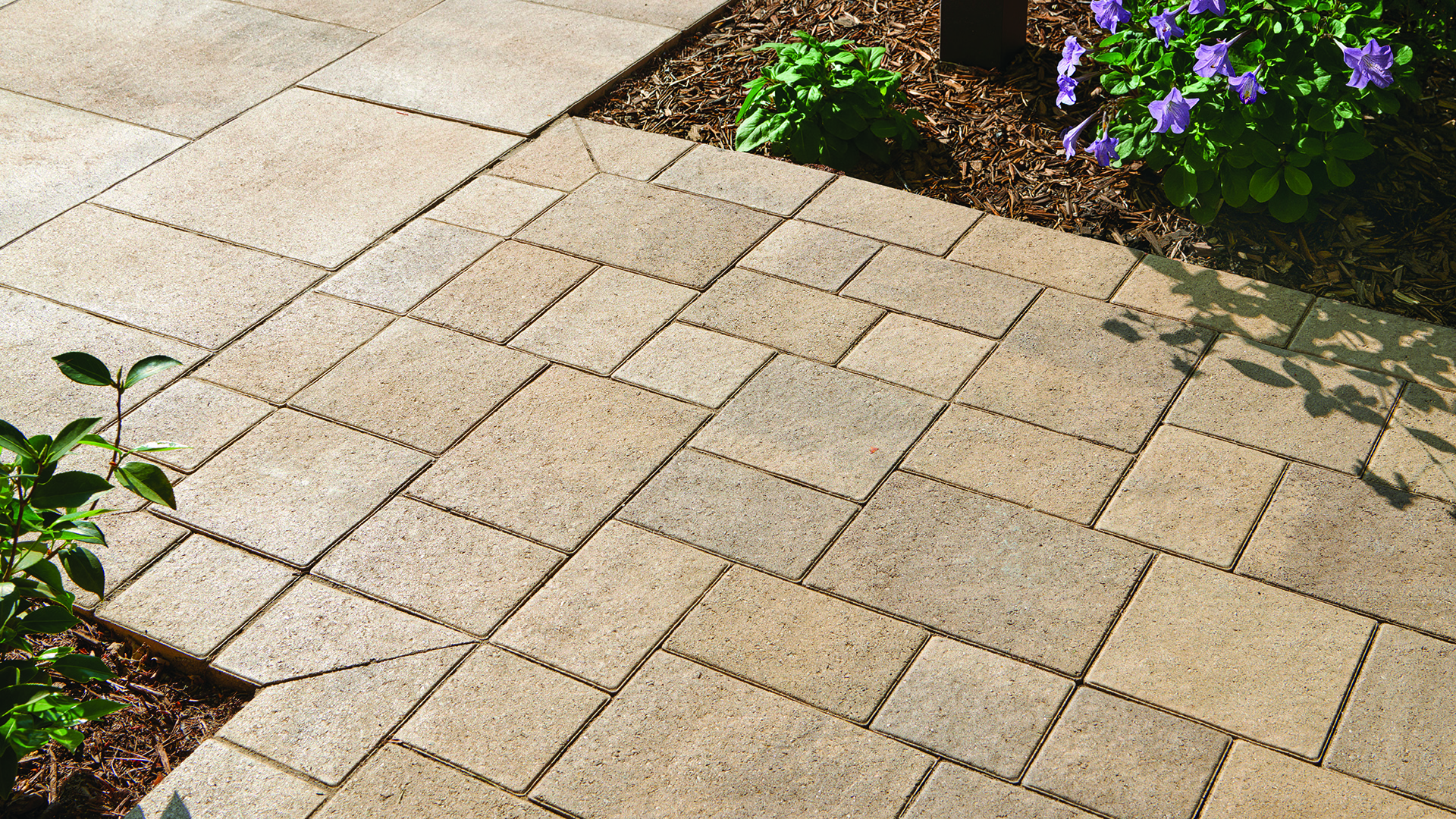 Close-up of a paved walkway featuring Porcellana 9-inch pavers in Light Sand-Gray-Stone installed in a random ashlar pattern in the foreground. Larger Porcellana 18-inch pavers in matching Light Sand-Gray-Stone are installed in a random ashlar pattern in the background, creating a coordinated transition between paving areas bordered by landscaped planting beds.