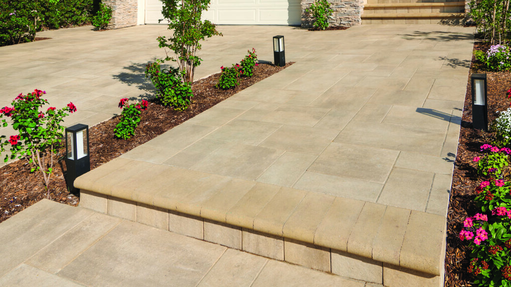 Entry walkway and driveway featuring Porcellana 18-inch pavers in Light Sand-Gray-Stone installed in a random ashlar pattern. The broad paved surface leads to a home's front entrance and garage, accented by landscaped planting beds with flowering shrubs. Steps are finished with 6x14 Bullnose units in Sand-Copper-Stone, providing a smooth transition between elevations and complementing the warm-toned porcelain paving.