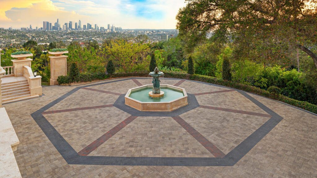 Porcellana 9-inch pavers in a Light Sand, Gray, and Stone color blend, installed in a random ashlar pattern, create a grand terrace surrounding a central fountain. Decorative banding of Antique Cobble I in Red, Brown, and Charcoal and Courtyard 6x9 in Charcoal forms a geometric radial design, with panoramic city skyline views framed by mature trees and landscaped gardens.