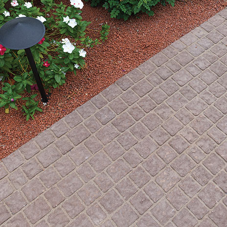 Close-up view of a garden pathway paved with 70mm Torino pavers in Mocha, installed in a random running bond pattern. The walkway is bordered by red decorative gravel, flowering plants, and landscape lighting, highlighting the textured surface and warm, natural color tones of the pavers.